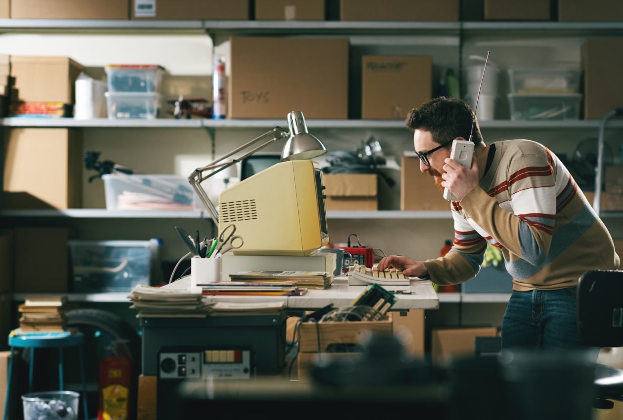 Vintage style nerd using an old computer in the garage and having a phone call with a cordless telephone